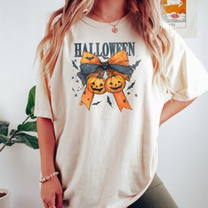 Puede incluir: Camiseta color crema con un dise&ntilde;o tem&aacute;tico de Halloween. El dise&ntilde;o presenta un lazo con cintas naranjas, un lazo negro y dos calabazas talladas. La palabra "HALLOWEEN" est&aacute; impresa encima del lazo en una fuente azul oscuro. Murci&eacute;lagos y estrellas rodean el lazo.