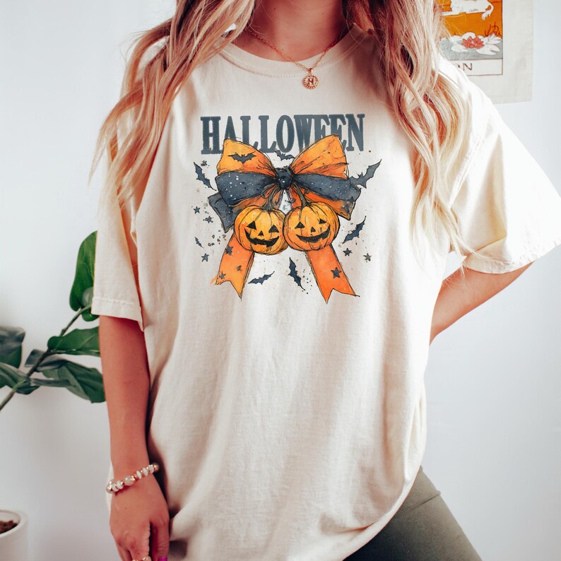 Puede incluir: Camiseta color crema con un dise&ntilde;o tem&aacute;tico de Halloween. El dise&ntilde;o presenta un lazo con cintas naranjas, un lazo negro y dos calabazas talladas. La palabra "HALLOWEEN" est&aacute; impresa encima del lazo en una fuente azul oscuro. Murci&eacute;lagos y estrellas rodean el lazo.