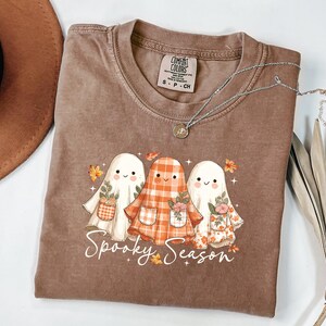 May include: A brown t-shirt with a graphic of three cartoon ghosts wearing floral and plaid outfits. The text "Spooky Season" is written in a script font below the ghosts. A silver necklace with a pendant is on the shirt.