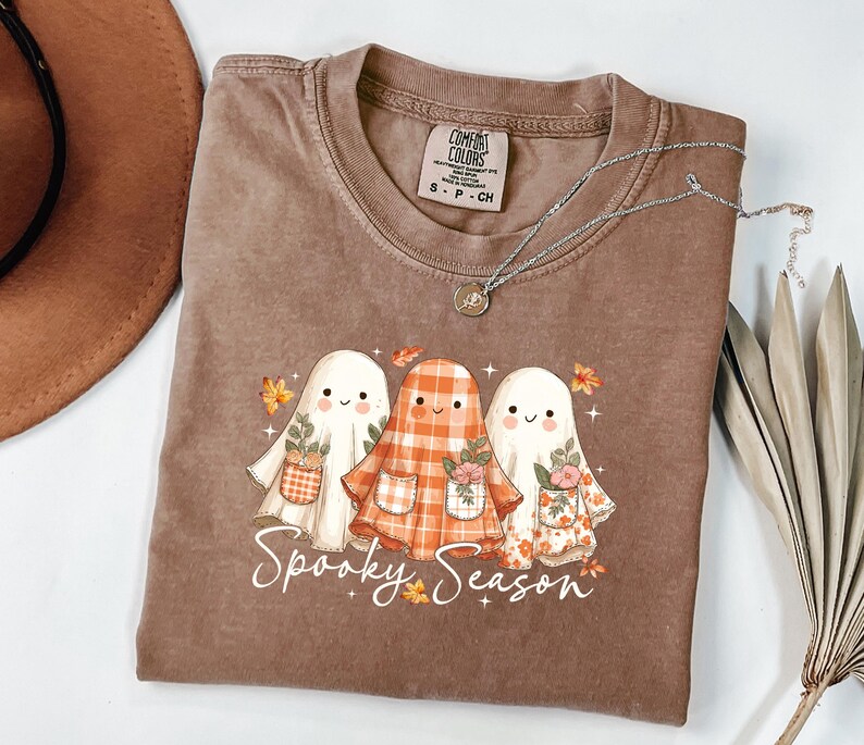May include: A brown t-shirt with a graphic of three cartoon ghosts wearing floral and plaid outfits. The text "Spooky Season" is written in a script font below the ghosts. A silver necklace with a pendant is on the shirt.