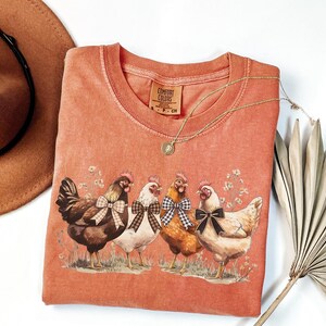 May include: Coral-colored Comfort Colors t-shirt featuring a graphic of four chickens with bow ties. The chickens are brown, white, and tan, standing on grass with floral accents. A gold necklace and a brown hat are also visible.
