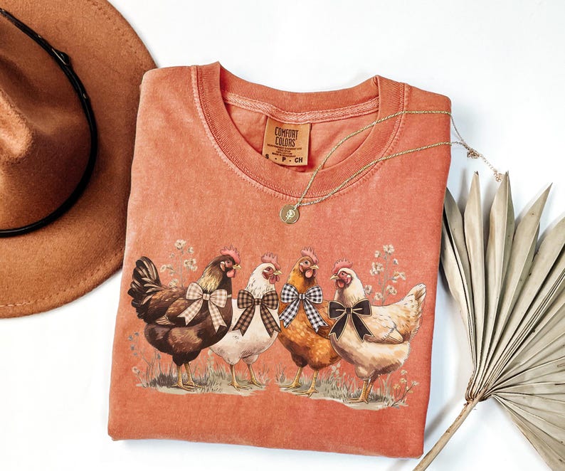 May include: Coral-colored Comfort Colors t-shirt featuring a graphic of four chickens with bow ties. The chickens are brown, white, and tan, standing on grass with floral accents. A gold necklace and a brown hat are also visible.