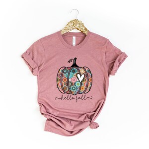 Puede incluir: Una camiseta color malva con una calabaza con estampado floral y el texto "hello fall" en letra negra.