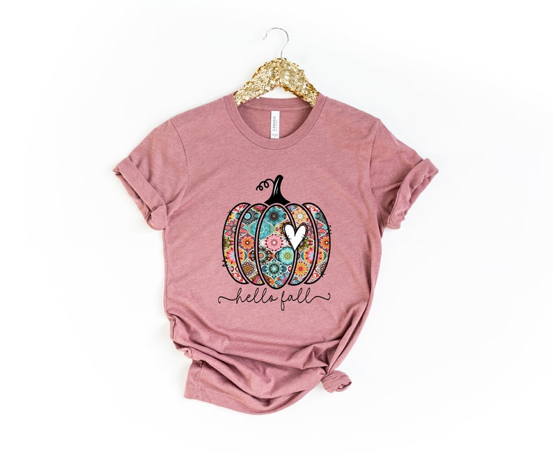 Puede incluir: Una camiseta color malva con una calabaza con estampado floral y el texto "hello fall" en letra negra.