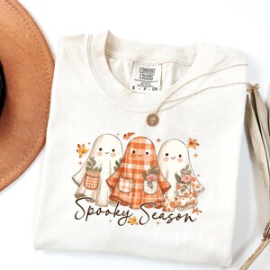 May include: Cream-colored t-shirt featuring three cartoon ghosts with floral and plaid patterns. The ghosts are holding small flower pots and are surrounded by autumn leaves. The text "Spooky Season" is written in a script font.
