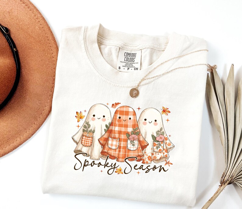 May include: Cream-colored t-shirt featuring three cartoon ghosts with floral and plaid patterns. The ghosts are holding small flower pots and are surrounded by autumn leaves. The text "Spooky Season" is written in a script font.