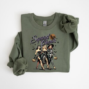May include: Green sweatshirt with a graphic of three women dressed as witches for Halloween. The text "Squad Goals" is printed on the sweatshirt.