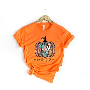 Puede incluir: Camiseta naranja con un gr&aacute;fico en blanco y negro de una calabaza con un coraz&oacute;n. La calabaza est&aacute; decorada con coloridos patrones florales. El texto "hello fall" est&aacute; escrito debajo de la calabaza.