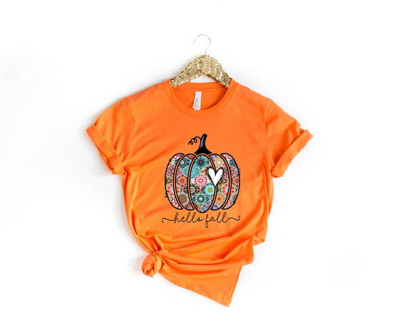 Puede incluir: Camiseta naranja con un gr&aacute;fico en blanco y negro de una calabaza con un coraz&oacute;n. La calabaza est&aacute; decorada con coloridos patrones florales. El texto "hello fall" est&aacute; escrito debajo de la calabaza.
