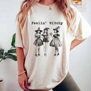May include: A white t-shirt with the text "Feelin' Witchy" and a graphic of three witches holding pumpkins and cups.