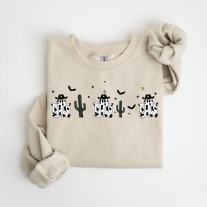 May include: Beige crewneck sweatshirt featuring a Halloween-themed design. The graphic includes three ghost-cowboy figures with black hats and cow-print bodies, along with cacti, bats, and stars. The sweatshirt has rolled sleeves.