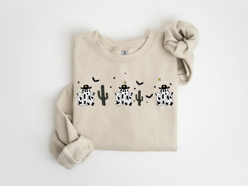 May include: Beige crewneck sweatshirt featuring a Halloween-themed design. The graphic includes three ghost-cowboy figures with black hats and cow-print bodies, along with cacti, bats, and stars. The sweatshirt has rolled sleeves.