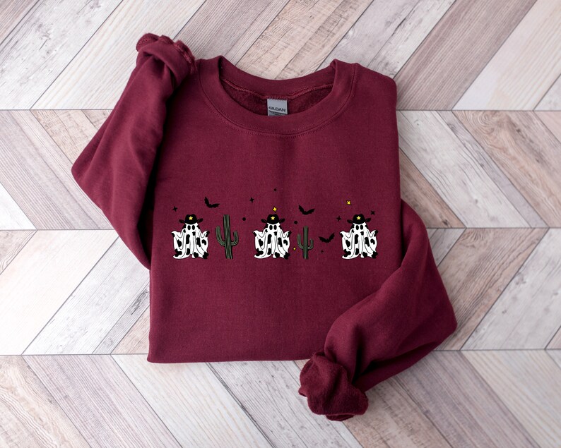 May include: A maroon sweatshirt featuring a Halloween-themed design. The design includes two ghosts wearing cowboy hats, a green cactus, and a bat. The ghosts have a black and white pattern. The sweatshirt is made of a soft material.