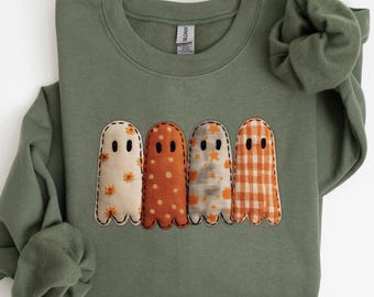 May include: An olive green sweatshirt with four appliqued ghost figures. Each ghost has a unique pattern: floral, polka dots, a grey and orange pattern, and orange gingham. The ghosts have black stitched eyes and a scalloped bottom edge.