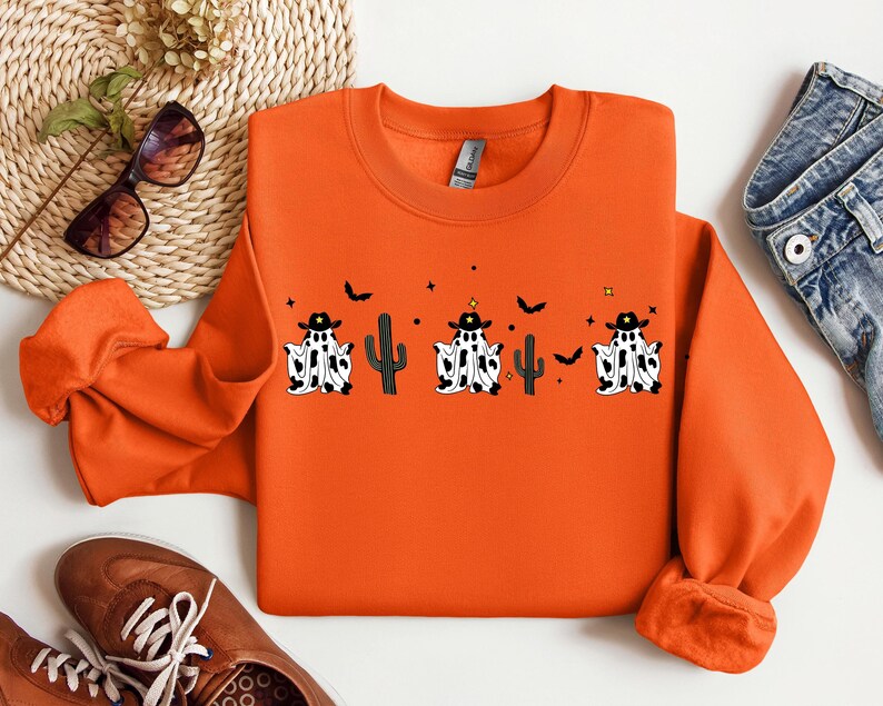 May include: Orange crewneck sweatshirt featuring a Halloween design. The design includes three ghost figures wearing cowboy hats, black and white cow print, and a cactus. Small black bats and stars are also included in the design.