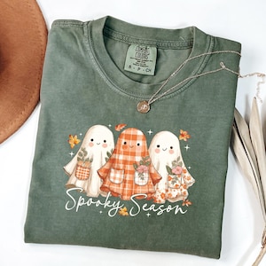 May include: Green Comfort Colors t-shirt featuring three cartoon ghosts with floral pockets and the text "Spooky Season." The ghosts are in white, orange plaid, and floral patterns. A silver necklace with a pendant is draped across the shirt.