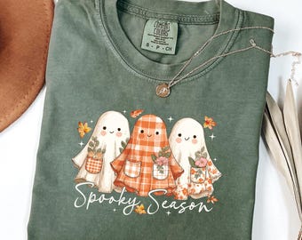 Könnte beinhalten: Grünes Comfort Colors T-Shirt mit drei Cartoon-Geistern mit Blumentaschen und dem Text "Spooky Season". Die Geister sind in Weiß, orangefarbenem Karomuster und floralen Mustern gehalten. Eine silberne Halskette mit Anhänger liegt über dem Shirt.