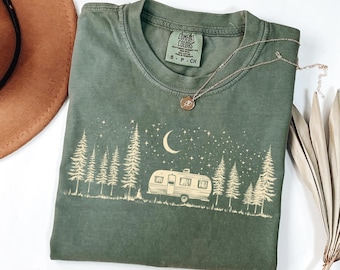 May include: An olive green Comfort Colors t-shirt featuring a camping scene with a trailer, trees, moon, and stars. A silver necklace with a pendant is draped across the shirt. A brown hat and dried plant are in the background.
