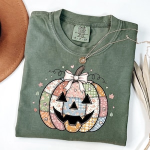 以下が含まれることがあります： パッチワークのかぼちゃのデザインが施された、グリーンのComfort Colors Tシャツ。かぼちゃは、花柄、チェック柄、無地の生地パターン、リボン、ジャック・オー・ランタンの顔が特徴です。ペンダント付きのゴールドネックレスがシャツの上にかけられています。