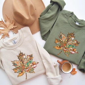 Könnte beinhalten: Zwei Sweatshirts, eines in Creme und eines in Olivgrün, jeweils mit einem Ahornblatt-Design. Das Blatt ist ein Patchwork aus herbstlichen Mustern und Farben. Ein beiger Hut und eine Tasse Kaffee sind ebenfalls im Bild.