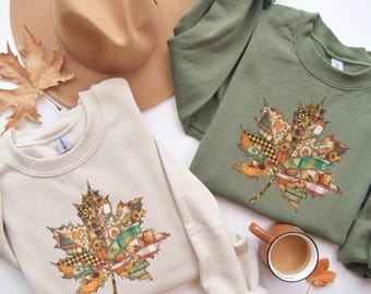 Puede incluir: Dos sudaderas, una crema y otra verde oliva, cada una con un diseño de hoja de arce. La hoja es un mosaico de patrones y colores otoñales. Un sombrero beige y una taza de café también están en la imagen.