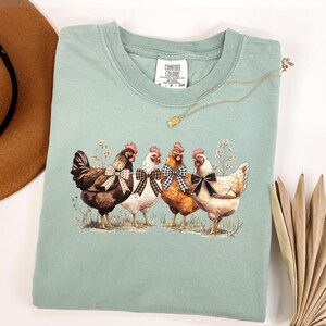 May include: A light green Comfort Colors t-shirt featuring a graphic of four chickens with bow ties. The chickens are brown, white, and orange, standing in a field of wildflowers. A gold necklace and a brown hat are also in the image.