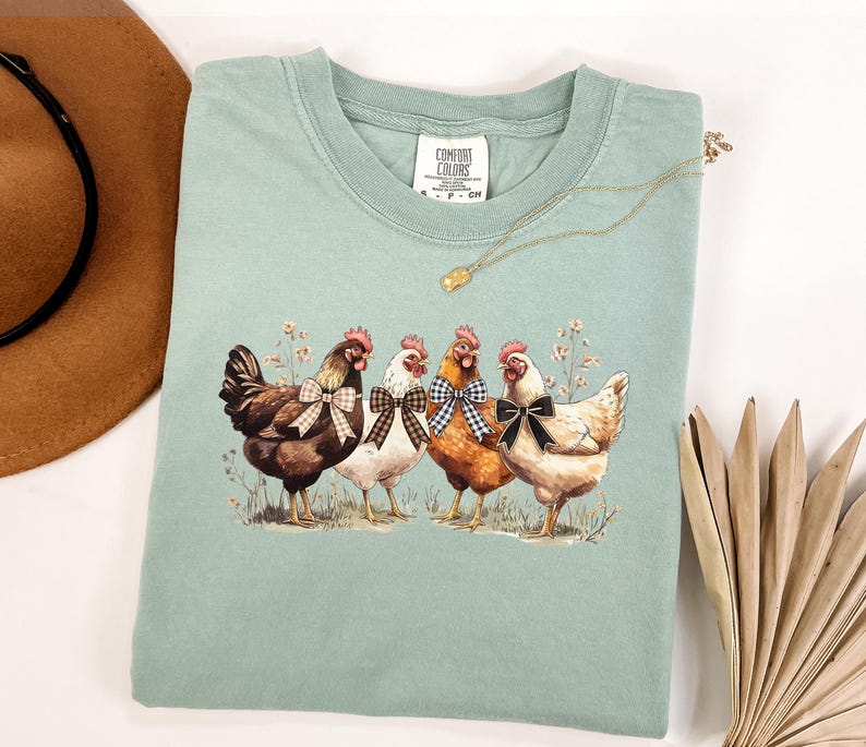 May include: A light green Comfort Colors t-shirt featuring a graphic of four chickens with bow ties. The chickens are brown, white, and orange, standing in a field of wildflowers. A gold necklace and a brown hat are also in the image.