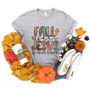 May include: A gray t-shirt with the text "Fall + Jesus he never leaves" in colorful letters with a leopard print design. The shirt is surrounded by fall-themed items like pumpkins, leaves, and a plaid scarf.
