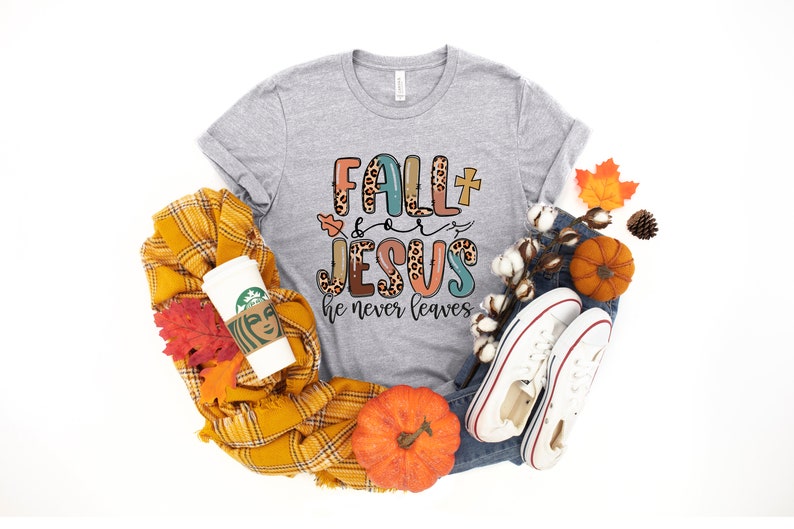 May include: A gray t-shirt with the text "Fall + Jesus he never leaves" in colorful letters with a leopard print design. The shirt is surrounded by fall-themed items like pumpkins, leaves, and a plaid scarf.