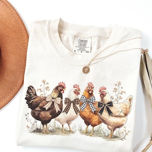 May include: Cream-colored Comfort Colors t-shirt featuring a watercolor illustration of four chickens with bowties. The chickens are brown, white, and orange, standing in a field of wildflowers. A gold necklace and a brown hat are also in the image.