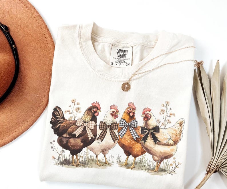 May include: Cream-colored Comfort Colors t-shirt featuring a watercolor illustration of four chickens with bowties. The chickens are brown, white, and orange, standing in a field of wildflowers. A gold necklace and a brown hat are also in the image.