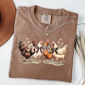 May include: A brown Comfort Colors t-shirt featuring a graphic of four chickens with bowties. The chickens are standing in a field of grass and flowers. A silver necklace with a pendant is draped across the shirt.