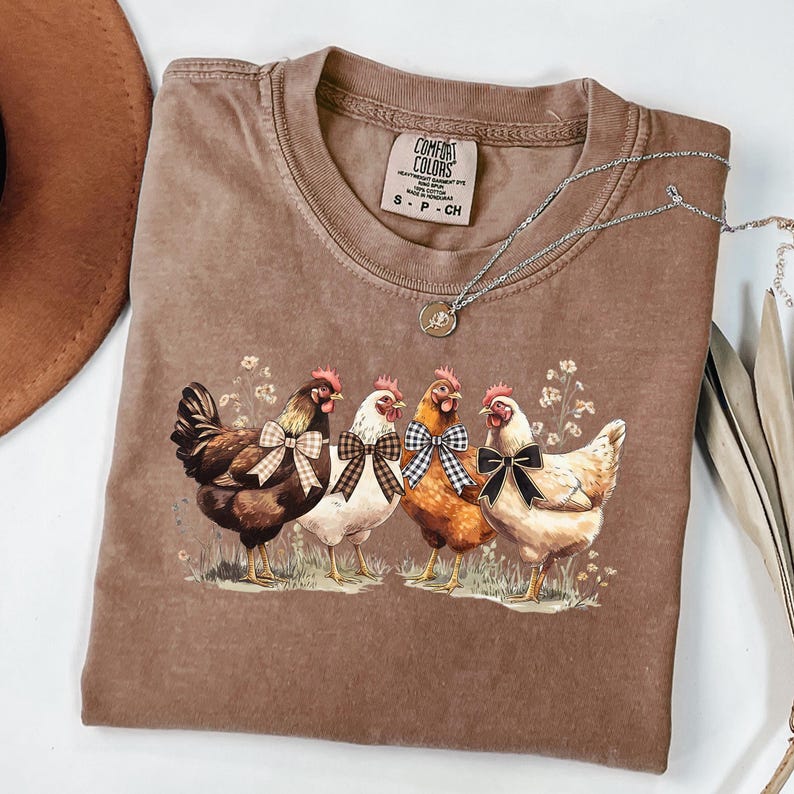 May include: A brown Comfort Colors t-shirt featuring a graphic of four chickens with bowties. The chickens are standing in a field of grass and flowers. A silver necklace with a pendant is draped across the shirt.