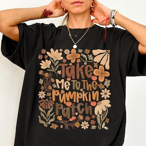 以下が含まれることがあります： 黒いTシャツ。茶色、オレンジ、白で「Take Me To The Pumpkin Patch」の文字と花柄のデザイン。秋色の花と葉が描かれています。