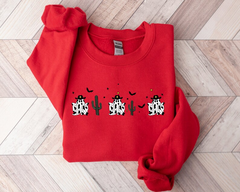 May include: A red sweatshirt featuring a Halloween-themed design. The design includes three ghosts wearing cowboy hats and cow-print outfits, a cactus, bats, and stars. The sweatshirt is made of a soft material.