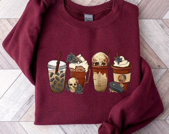 May include: Burgundy sweatshirt featuring a graphic of four Halloween-themed coffee drinks. Each drink has a skull or coffin design, with details like whipped cream, roses, and a coffin. The sweatshirt is folded on a light wood-grain surface.