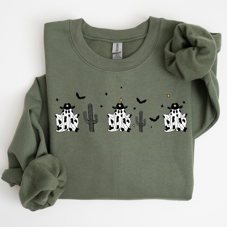 May include: Olive green sweatshirt featuring a graphic of three ghost figures wearing cowboy hats with black and white cow print. The design includes black bats, stars, and cacti. The sweatshirt has a crew neck and long sleeves.