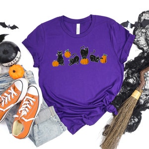 May include: A purple t-shirt with a Halloween-themed design. The graphic depicts black cats and orange pumpkins. The cats are in different poses. The shirt is styled with orange trainers and blue jeans.