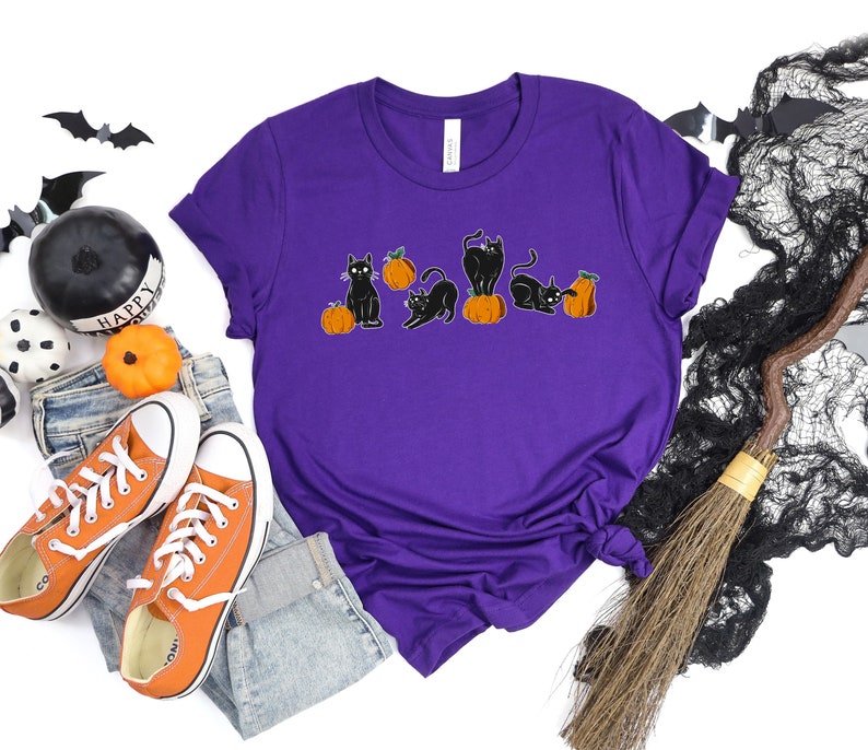 May include: A purple t-shirt with a Halloween-themed design. The graphic depicts black cats and orange pumpkins. The cats are in different poses. The shirt is styled with orange trainers and blue jeans.