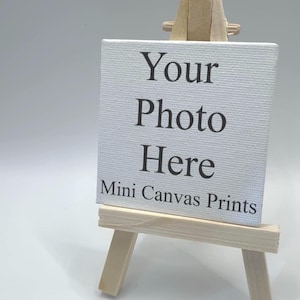 May include: A small square white canvas print resting on a light-colored wooden tripod easel. The canvas features the black printed text "Your Photo Here" and "Mini Canvas Prints" centered on the front surface against a plain white background.