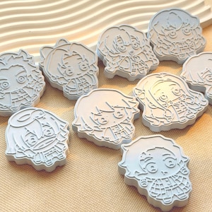 May include: A collection of nine light gray, flat-surfaced cookie cutters featuring embossed chibi-style anime character designs. Each cutter has a distinct outline and detailed facial features, including large eyes and hair, molded into the plastic.