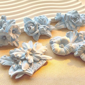 Op de afbeelding: Vijf lichtblauwe bloemenornamenten met gebeeldhouwde rozen, hibiscus en andere bloesems. Eén stuk toont het woord Hawaii in reliëfletters. Deze decoratieve items hebben gedetailleerde bloemblaadjes en bladeren met een matte afwerking.