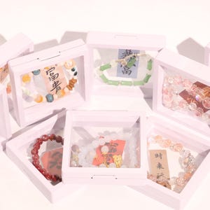 Puede incluir: Una colección de pulseras exhibidas en cajas rosas. Las pulseras presentan diversas cuentas en colores como rojo, naranja, amarillo, verde y blanco. Algunas cajas contienen pequeñas tarjetas con caracteres chinos.