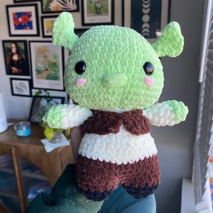 May include: A green crocheted plush toy with a brown vest and black shoes. The toy has large eyes and a small pink nose.