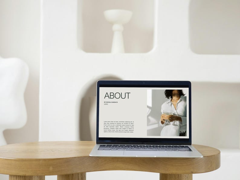 Pu&ograve; includere: Un laptop che mostra una pagina web con la parola "ABOUT". Il laptop &egrave; su un tavolo rotondo di legno. Lo sfondo &egrave; una parete bianca con nicchie decorative.