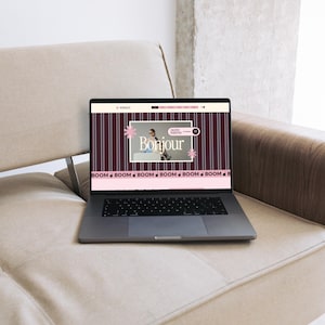 May include: A dark gray laptop displaying a website with a dark purple background featuring vertical white stripes. The screen shows the text Bonjour, a small photograph, and a horizontal pink banner at the bottom with the repeated word BOOM.