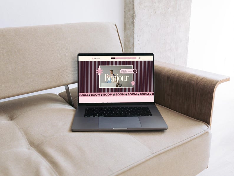 May include: A dark gray laptop displaying a website with a dark purple background featuring vertical white stripes. The screen shows the text Bonjour, a small photograph, and a horizontal pink banner at the bottom with the repeated word BOOM.
