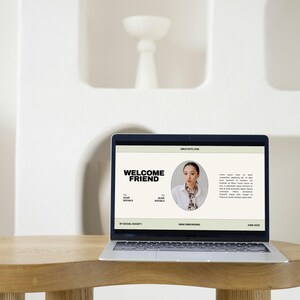 May include: A laptop displaying a website with the text "WELCOME FRIEND" in bold black letters. The website features a photo of a person and additional text. The laptop sits on a round wooden table.