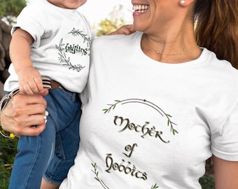 May include: A mother and child wearing white t-shirts with a green leafy wreath design. The child's shirt says "Halfling" and the mother's shirt says "Mother of Hobbits".