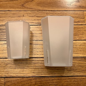 May include: Two frosted, geometric-shaped planters. The planters are made of a translucent material, with a rectangular shape and a slightly angled top. The planters are empty and sit on a wooden surface.